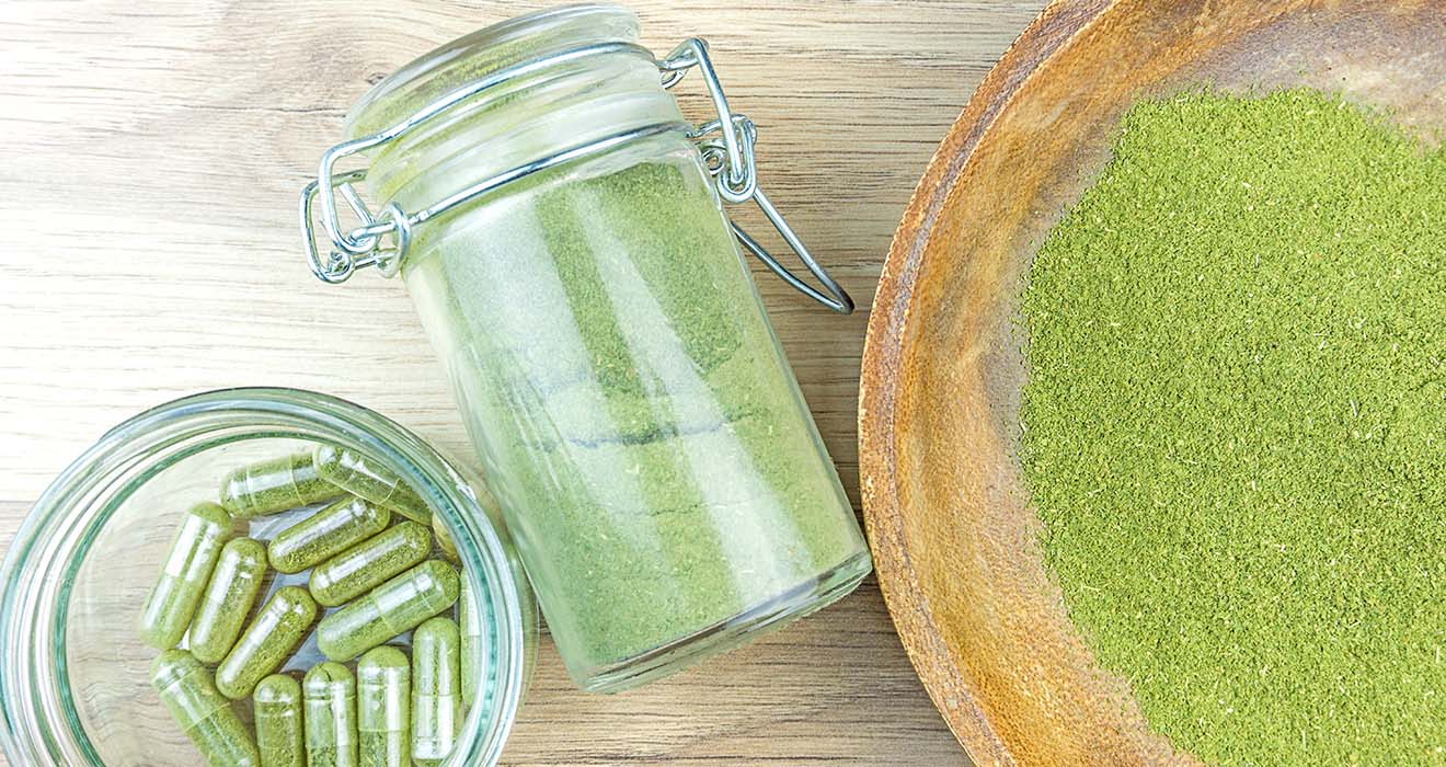 Green powder and capsules arranged on a wooden surface, showcasing health supplements in glass jars and a wooden bowl.