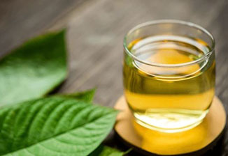 A clear glass filled with yellow-green liquid sits on a wooden surface, surrounded by fresh green leaves.