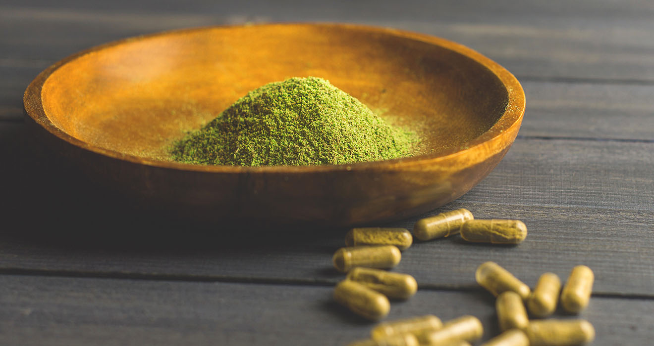 A wooden bowl filled with green powder sits on a dark surface, surrounded by scattered green capsules.