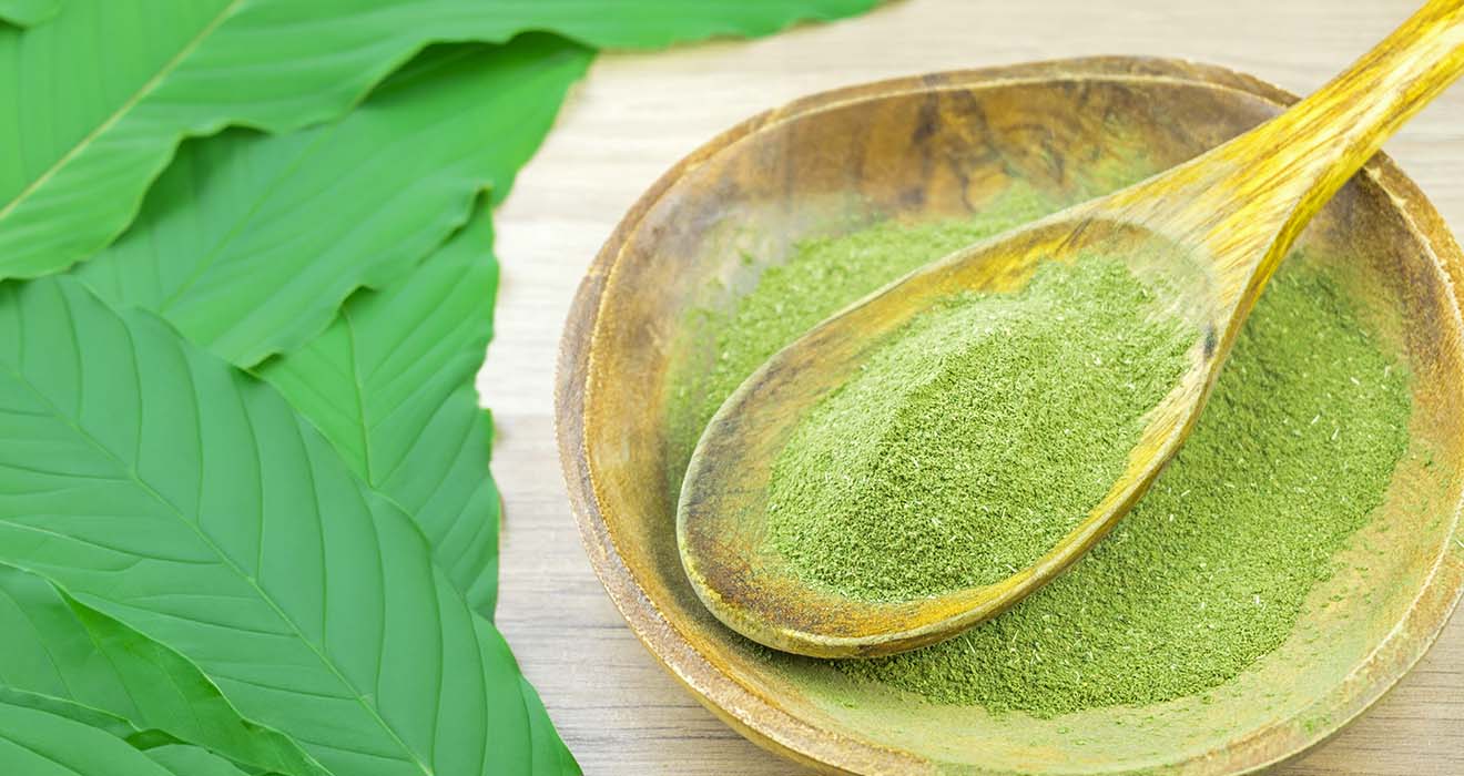 A wooden bowl filled with green powder sits beside large green leaves on a wooden surface, suggesting a natural ingredient.